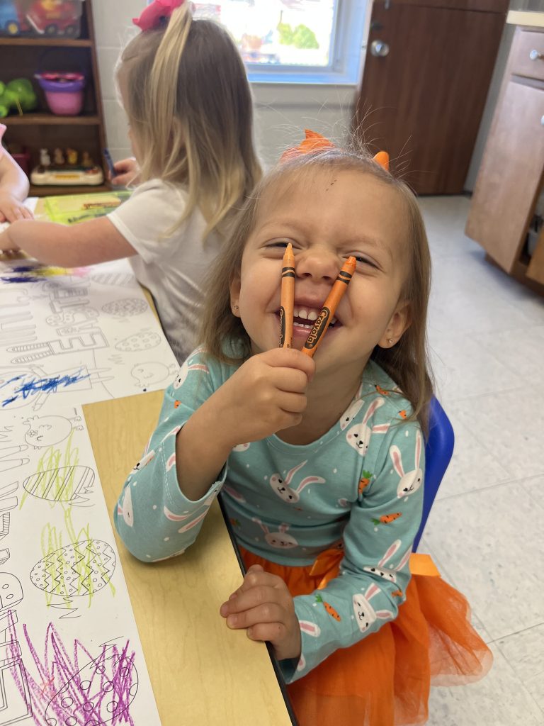 A child grins at the camera while making a silly face and holding up crayons.