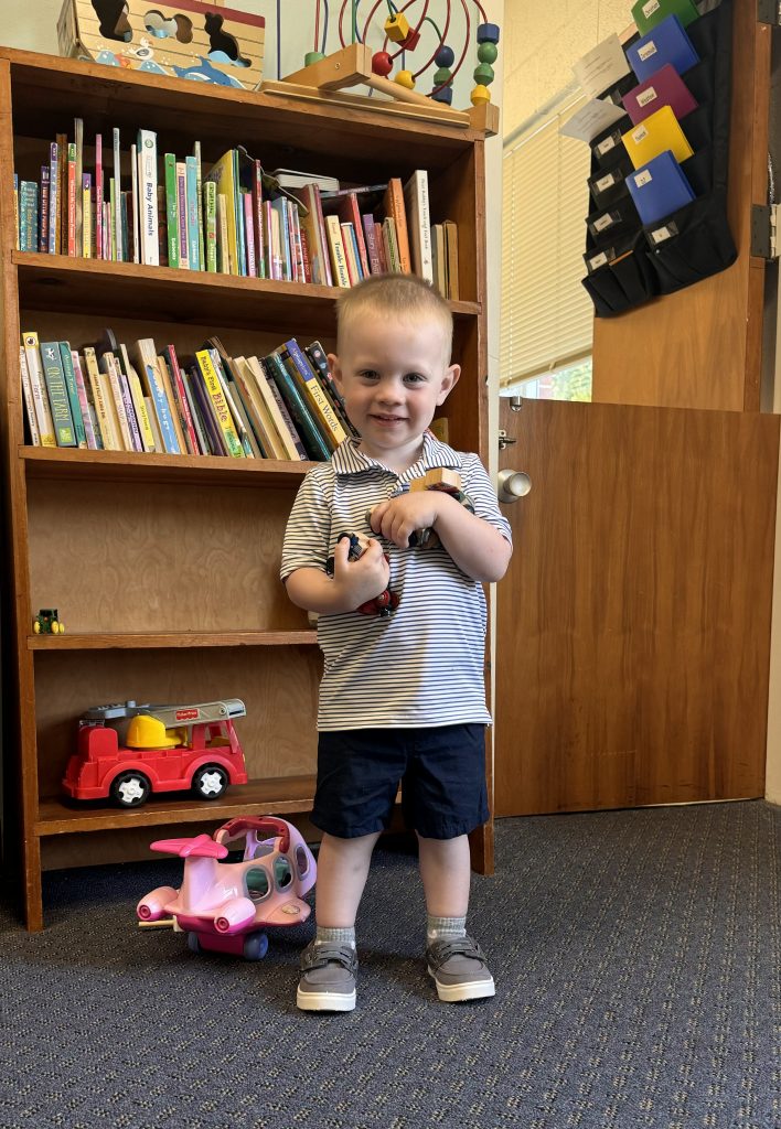 A child holding toys stands in front of a bookcase.