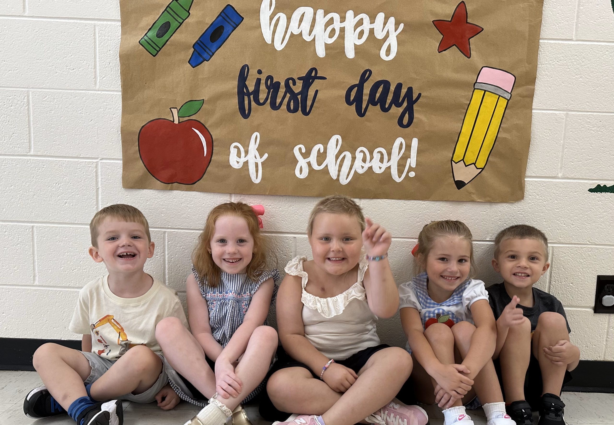 Children smile at the camera. A sign above them reads Happy First Day of School